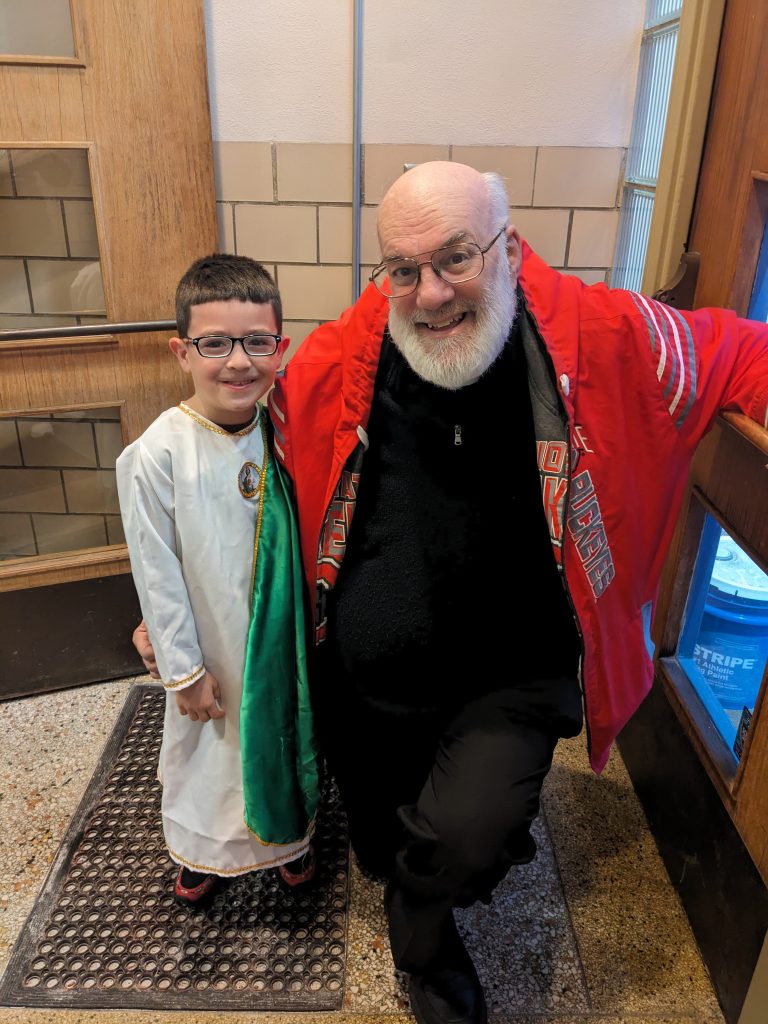 Young student dressed as a priest in appropriate attire, next to Fr. Paul, kneeled down. Both smiling, posing for picture